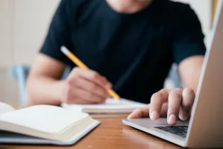 A person sits with pencil in hand at a laptop computer, scribbling and typing.
